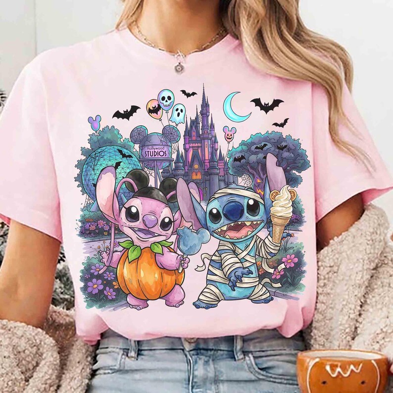 May include: A light pink t-shirt featuring a graphic print of two blue alien characters. One character is inside a pumpkin, and the other is wrapped in bandages like a mummy holding an ice cream cone. The background shows a castle and park.