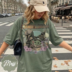 Puede incluir: Camiseta verde musgo con un dise&ntilde;o de The Mandalorian y Grogu rodeados de flores. El texto "MANDALORIAN" est&aacute; impreso en naranja y marr&oacute;n sobre el dise&ntilde;o. La camiseta es de corte holgado.
