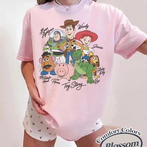 May include: A pale pink t-shirt with a graphic of Toy Story characters, including Woody and Buzz Lightyear. The shirt has the text "Toy Story" and "Comfort Colors Blossom".