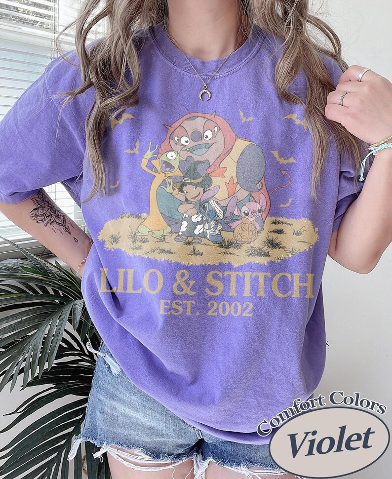 May include: A violet Comfort Colors t-shirt featuring a graphic of characters from Lilo & Stitch, with the text "Lilo & Stitch EST. 2002". The design includes Stitch, Lilo, and other characters in a Halloween theme.