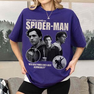 May include: A purple t-shirt featuring Tom Holland as Spider-Man. The design includes multiple images of Holland, the Spider-Man logo, and the quote "WITH GREAT POWER COMES GREAT RESPONSIBILITY".