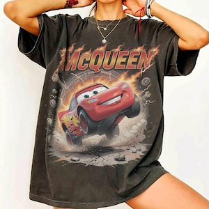 Op de afbeelding: Donkergrijze t-shirt met een afbeelding van Bliksem McQueen uit de film Cars. Het ontwerp bevat de naam "McQUEEN" in een vlammende lettertype, met het nummer 95 op de auto. Het t-shirt heeft een vintage, verweerde uitstraling.