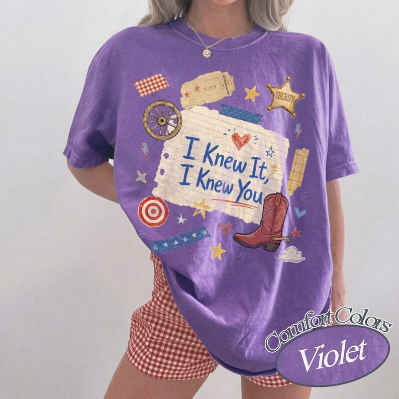 May include: A violet short-sleeve t-shirt featuring a central graphic of a lined notebook paper with the text I Knew It, I Knew You. The design includes a red cowboy boot, a sheriff star, a wagon wheel, stars, and tape strips.