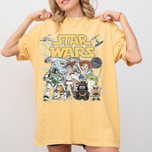 Puede incluir: Camiseta amarilla mostaza con un gr&aacute;fico de personajes de Star Wars, incluyendo a Darth Vader, Woody y Buzz Lightyear. Las palabras "STAR WARS" est&aacute;n impresas en amarillo sobre los personajes. La camiseta es de estilo casual.