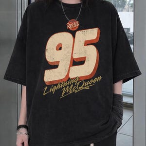 Puede incluir: Camiseta negra con el n&uacute;mero "95" en crema con contorno rojo, y las palabras "Lightning McQueen" en escritura amarilla. La camiseta tambi&eacute;n tiene la palabra "Rusteze" en un c&iacute;rculo rojo. La camiseta tiene mangas cortas.