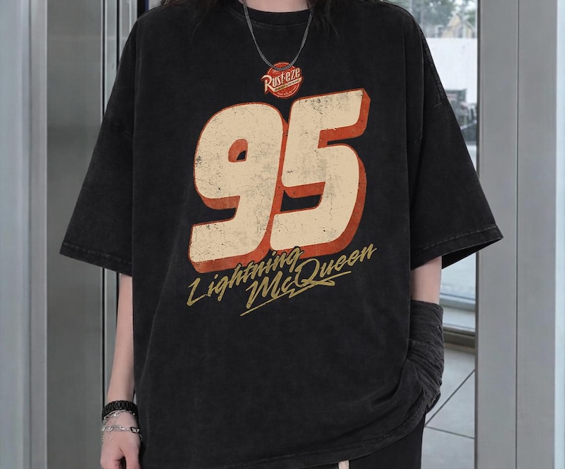 Puede incluir: Camiseta negra con el n&uacute;mero "95" en crema con contorno rojo, y las palabras "Lightning McQueen" en escritura amarilla. La camiseta tambi&eacute;n tiene la palabra "Rusteze" en un c&iacute;rculo rojo. La camiseta tiene mangas cortas.