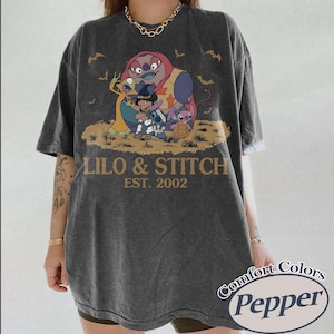 May include: Dark gray t-shirt featuring a colorful graphic of Lilo & Stitch characters with the text "LILO & STITCH EST. 2002". The shirt has a vintage look and the Comfort Colors Pepper logo.