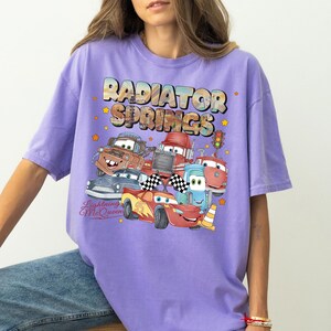 May include: A lilac-colored t-shirt featuring a graphic of characters from the movie Cars. The design includes the text "Radiator Springs" and "Lightning McQueen" with images of various car characters.