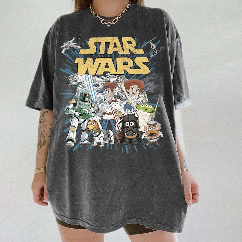 Puede incluir: Camiseta gris oscuro con un dise&ntilde;o de personajes de Star Wars, incluyendo Darth Vader, Woody y Buzz Lightyear. Las palabras "STAR WARS" est&aacute;n en letras amarillas. La camiseta tiene mangas cortas.