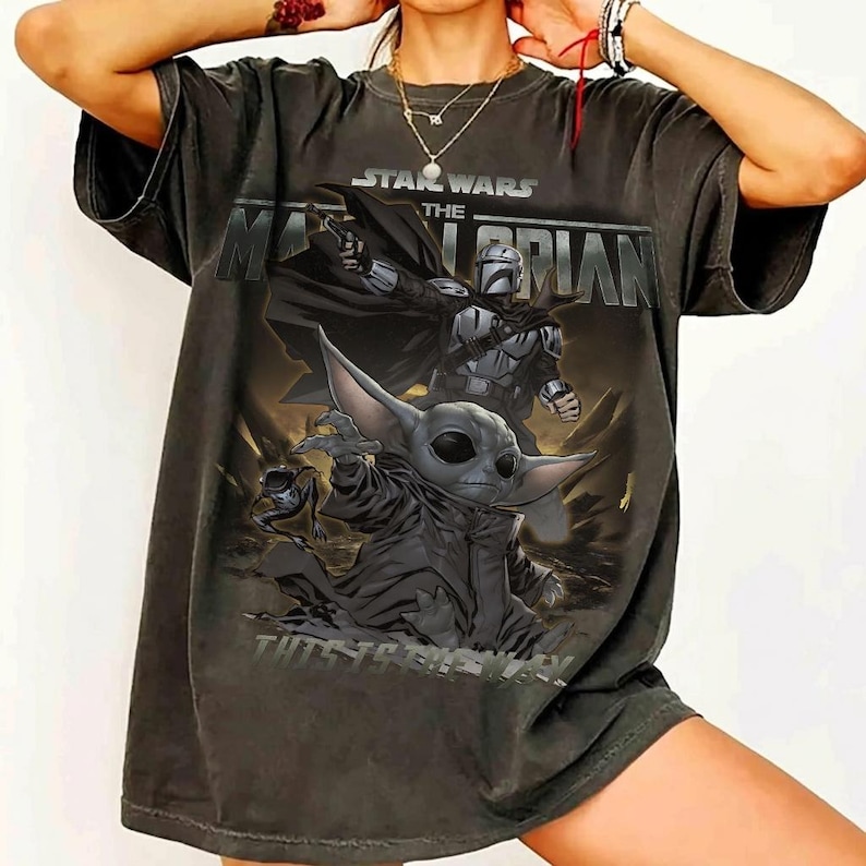 May include: Dark grey t-shirt with a graphic of The Mandalorian and Grogu from Star Wars. The design includes the text "STAR WARS THE MANDALORIAN" and "THIS IS THE WAY". The shirt has a vintage, distressed appearance.