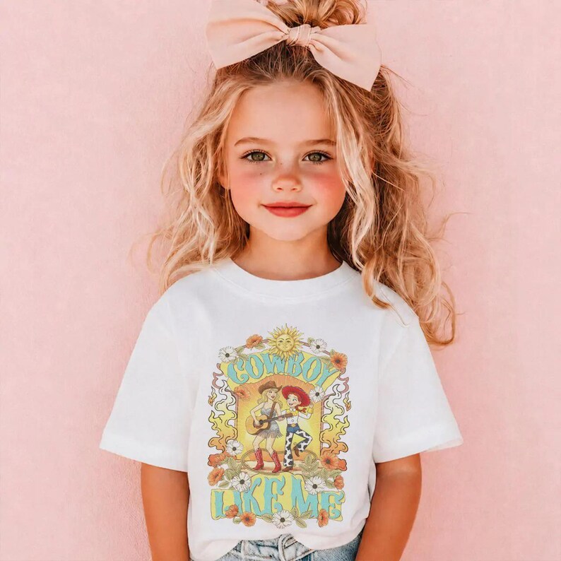 May include: A white short-sleeved t-shirt featuring a graphic print with the text COWBOY LIKE ME. The design includes illustrations of two figures, one playing a guitar, surrounded by yellow flames, orange flowers, and a sun icon.