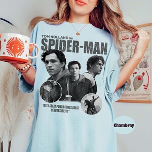 May include: Light blue t-shirt featuring Tom Holland as Spider-Man. The design includes images of the actor and the text "WITH GREAT POWER COMES GREAT RESPONSIBILITY". The shirt is a chambray color.