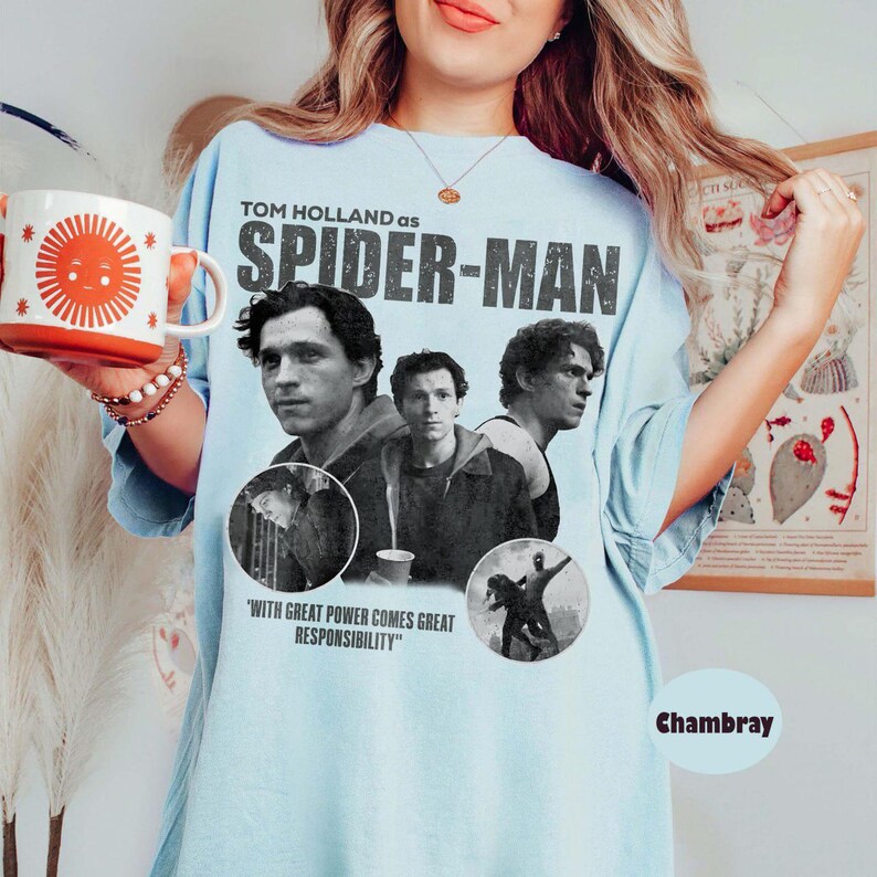 May include: Light blue t-shirt featuring Tom Holland as Spider-Man. The design includes images of the actor and the text "WITH GREAT POWER COMES GREAT RESPONSIBILITY". The shirt is a chambray color.