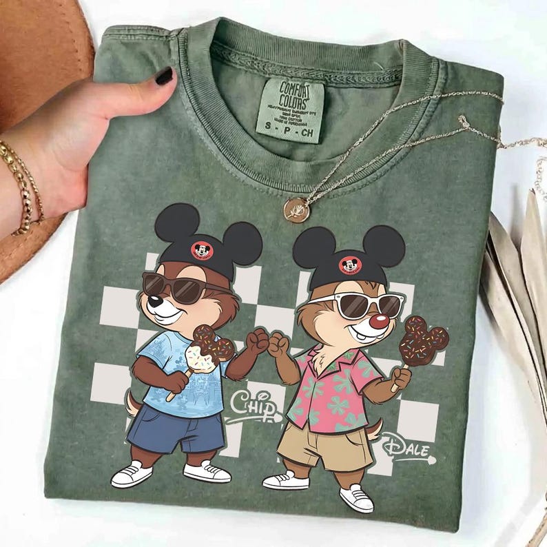 Pu&ograve; includere: T-shirt verde Comfort Colors con un'illustrazione di Chip e Dale con occhiali da sole e gelati al cioccolato. La maglietta ha uno sfondo a scacchi e le parole "Chip" e "Dale".