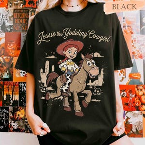 May include: A black short-sleeve t-shirt featuring a graphic print of Jessie the Yodeling Cowgirl riding a horse. The design includes the text Jessie the Yodeling Cowgirl in a cursive font, with desert elements like cacti and skulls in the background.