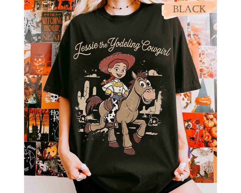 May include: A black short-sleeve t-shirt featuring a graphic print of Jessie the Yodeling Cowgirl riding a horse. The design includes the text Jessie the Yodeling Cowgirl in a cursive font, with desert elements like cacti and skulls in the background.
