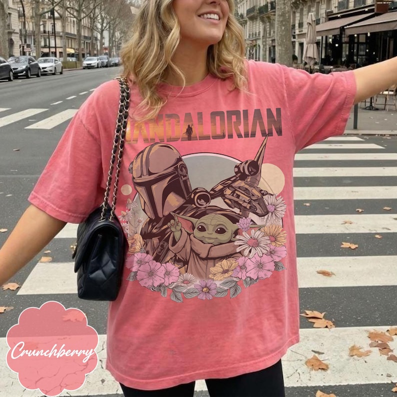 Puede incluir: Camiseta de color coral con un gr&aacute;fico de The Mandalorian, Grogu y una nave espacial, rodeados de flores. La palabra "MANDALORIAN" est&aacute; impresa en la parte superior en un estilo vintage. La camiseta tiene un corte holgado.
