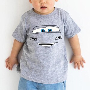 Op de afbeelding: Een grijs gem&ecirc;leerd T-shirt met korte mouwen en een grafische print van een cartoon autogezicht met grote witte voorruit-ogen, blauwe pupillen en een brede glimlachende mond, gecentreerd op de borst van het shirt met ronde hals.
