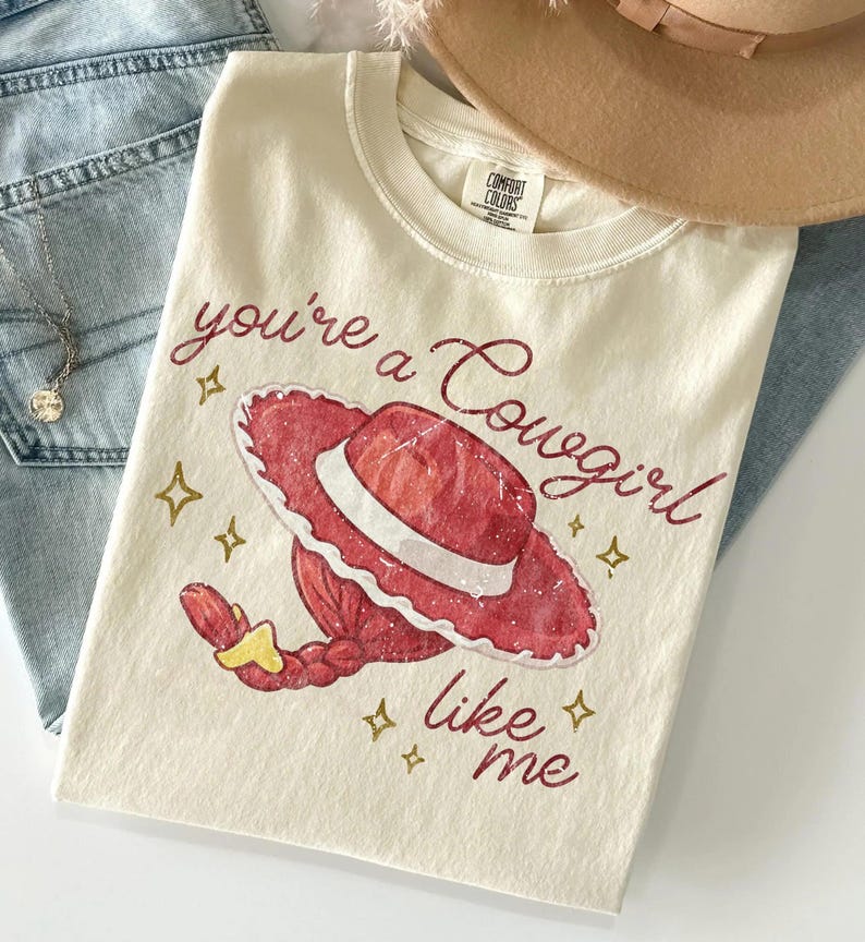 Puede incluir: Una camiseta de manga corta color crema con un gr&aacute;fico de un sombrero de vaquera rojo y un peinado trenzado. El texto you're a Cowgirl like me aparece en una fuente cursiva de color rojo oscuro, con peque&ntilde;as estrellas doradas impresas.