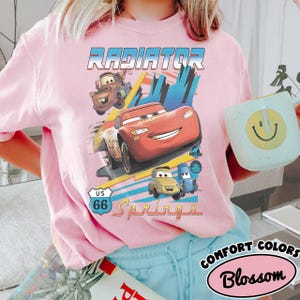 May include: A light pink t-shirt featuring a graphic print of characters from the movie Cars, including Lightning McQueen, Mater, Luigi, and Guido. The text Radiator Springs and a US 66 road sign are printed above and below the colorful car illustrations.