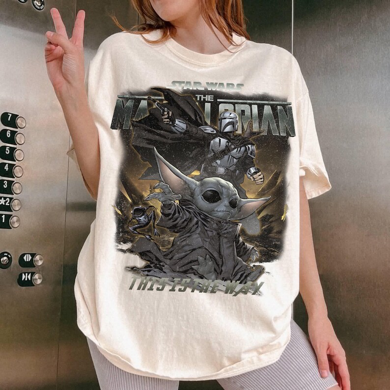 May include: A cream-coloured t-shirt with a graphic from the Star Wars series, "The Mandalorian." The design includes the text "STAR WARS THE MANDALORIAN" above an image of the Mandalorian and Grogu, with the phrase "THIS IS THE WAY" below.