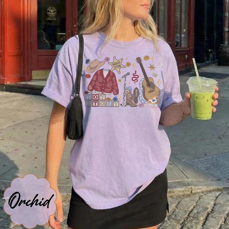 May include: A light purple short-sleeve t-shirt featuring a graphic print of a cowboy hat, a paisley cardigan, a sheriff star, a microphone, a guitar, and cowboy boots. The text "TOY STORY" is printed in block letters alongside the graphic elements.