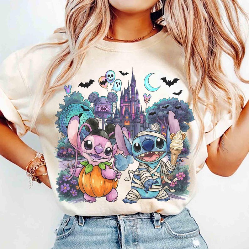 May include: A cream-colored t-shirt featuring a graphic print of two cartoon characters, one dressed as a pumpkin and the other as a mummy, standing in front of a theme park castle, with balloons, bats, and a crescent moon in the background.