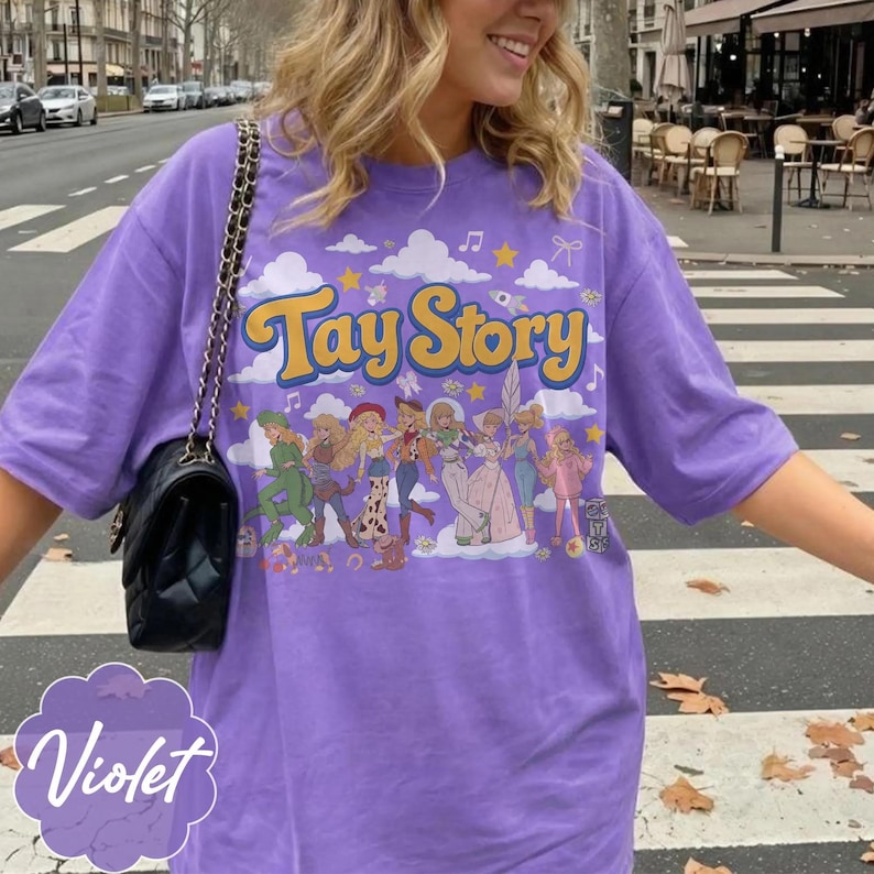May include: A violet short-sleeved t-shirt featuring the yellow text Tay Story above a graphic illustration of multiple figures in various outfits, surrounded by clouds, stars, and musical notes, paired with a black quilted shoulder bag.