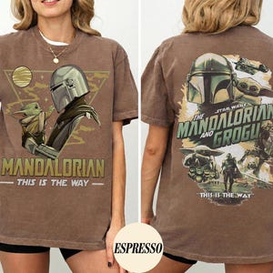K&ouml;nnte beinhalten: Braunes T-Shirt mit einer Grafik von The Mandalorian und Grogu. Das Frontdesign enth&auml;lt den Text "MANDALORIAN" und "THIS IS THE WAY". Das R&uuml;ckendesign zeigt den Text "STAR WARS THE MANDALORIAN AND GROGU" und "THIS IS THE WAY".