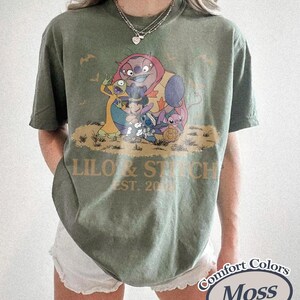 May include: Olive green t-shirt featuring a graphic of characters from Lilo & Stitch, with the text "Lilo & Stitch EST. 2002". The shirt is a Comfort Colors brand.