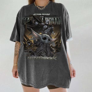May include: Dark grey t-shirt with a Star Wars "The Mandalorian" graphic. The design shows the Mandalorian and Grogu, with the text "This is the way." The shirt has a vintage, washed appearance.