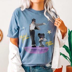 Puede incluir: Camiseta azul con un gr&aacute;fico de Michael Jackson y Mickey Mouse. La ilustraci&oacute;n muestra a Michael Jackson con traje y sombrero negros, y a Mickey Mouse con un atuendo similar. Las palabras "Michael Jackson" y "Mickey Mouse" tambi&eacute;n est&aacute;n en la camiseta.
