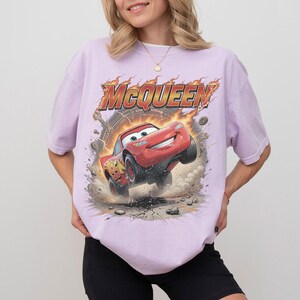 Op de afbeelding: Een lichtpaars T-shirt met een afbeelding van Bliksem McQueen uit de film Cars. De rode raceauto wordt in de sprong afgebeeld, met het woord "McQueen" erboven in een vurige lettertype. Het shirt is een comfortabele, casual stijl.