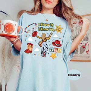 Peut inclure: Un t-shirt bleu clair avec une impression graphique portant le texte "I Knew It, I Knew You", un personnage de dessin anim&eacute; jouant de la guitare, un chapeau de cowboy rouge, des bottes de cowboy rouges, un bracelet de perles et des blocs "TS5".