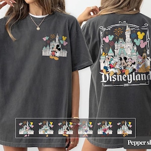 Pode incluir: T-shirt cinzenta escura com um design da Disneyland. A frente tem um pequeno gr&aacute;fico de Mickey e Minnie Mouse com um castelo e bal&otilde;es. A parte de tr&aacute;s exibe um gr&aacute;fico maior da Disneyland com personagens e fogos de artif&iacute;cio. O texto "Disneyland 1955" tamb&eacute;m &eacute; vis&iacute;vel.