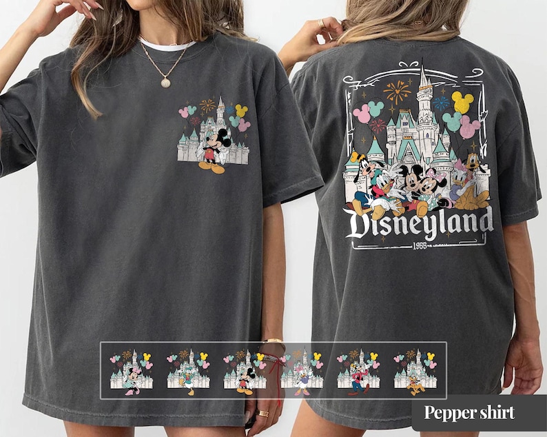 Pode incluir: T-shirt cinzenta escura com um design da Disneyland. A frente tem um pequeno gr&aacute;fico de Mickey e Minnie Mouse com um castelo e bal&otilde;es. A parte de tr&aacute;s exibe um gr&aacute;fico maior da Disneyland com personagens e fogos de artif&iacute;cio. O texto "Disneyland 1955" tamb&eacute;m &eacute; vis&iacute;vel.
