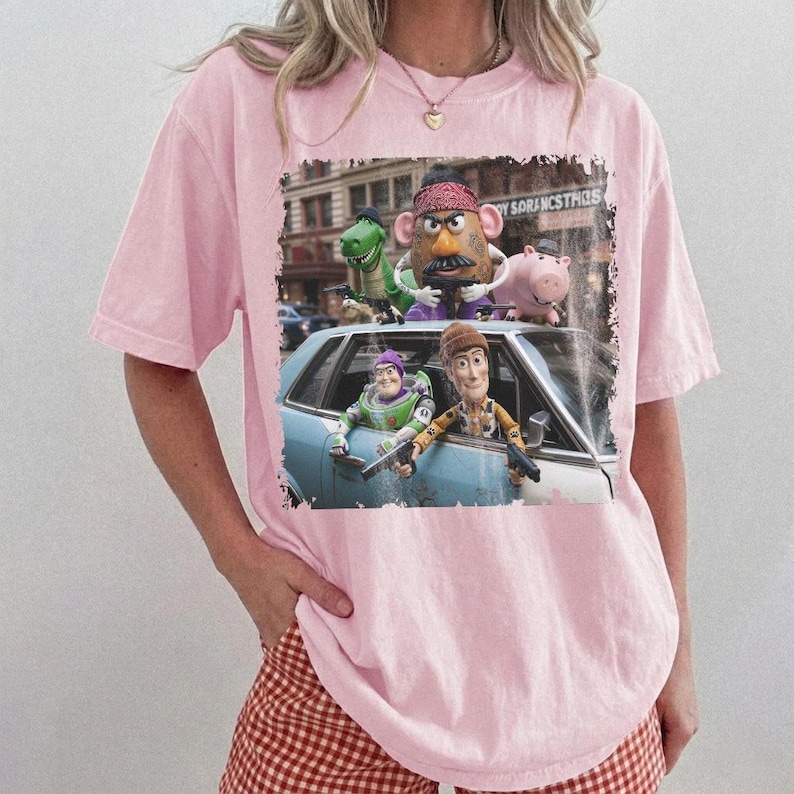 K&ouml;nnte beinhalten: Ein hellrosa T-Shirt mit kurzen &Auml;rmeln und einem Grafikdruck von Toy Story-Figuren, darunter Woody, Buzz Lightyear, Mr. Potato Head, Rex und Hamm in einem Auto. Der Druck zeigt die Charaktere in einer st&auml;dtischen Umgebung mit Rand.