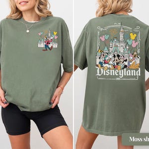 Pode incluir: Camiseta verde musgo com um gr&aacute;fico da Disneyland na frente e atr&aacute;s. O gr&aacute;fico frontal mostra Pateta com bal&otilde;es e um castelo. O gr&aacute;fico traseiro exibe Mickey Mouse, Minnie Mouse, Pato Donald e outros personagens com o logotipo da Disneyland.