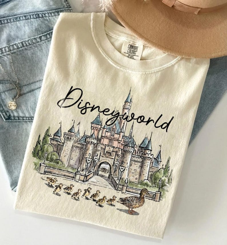 Puede incluir: Camiseta color crema con una ilustraci&oacute;n en acuarela de un castillo y la palabra "Disneyworld" en escritura negra. El gr&aacute;fico incluye una fila de patitos y un pato. Un sombrero beige y unos vaqueros azules est&aacute;n en el fondo.
