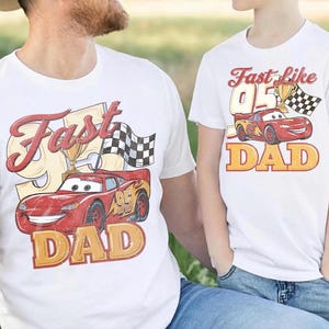 May include: Two white t-shirts with a red race car graphic. One shirt says "Fast Dad" with a checkered flag and trophy. The other says "Fast like Dad" with a similar design. Both shirts have a vintage style.