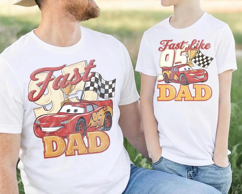 May include: Two white t-shirts with a red race car graphic. One shirt says "Fast Dad" with a checkered flag and trophy. The other says "Fast like Dad" with a similar design. Both shirts have a vintage style.