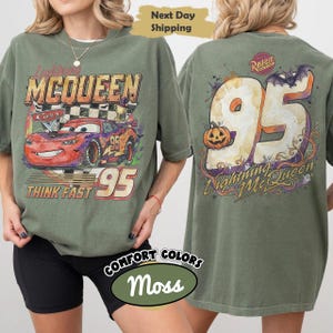 May include: A moss green t-shirt featuring a graphic of Lightning McQueen on the front with the text Lightning McQueen, Cars, and Think Fast 95. The back displays a large 95 with a pumpkin, a bat, and the text Rust-eze Lightning McQueen.