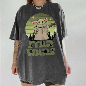 Op de afbeelding: Donkergrijs T-shirt met een Star Wars-afbeelding van Grogu. Het ontwerp bevat een cirkelvormige achtergrond met Grogu, een ruimteschip en de woorden "STAR WARS" in het groen. Het shirt heeft korte mouwen en een relaxte pasvorm.