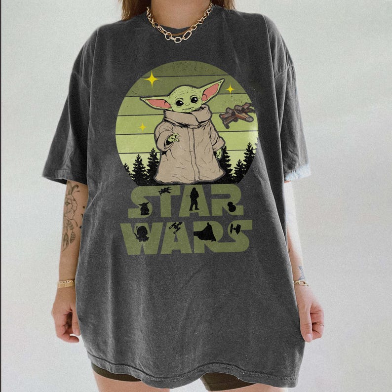Op de afbeelding: Donkergrijs T-shirt met een Star Wars-afbeelding van Grogu. Het ontwerp bevat een cirkelvormige achtergrond met Grogu, een ruimteschip en de woorden "STAR WARS" in het groen. Het shirt heeft korte mouwen en een relaxte pasvorm.