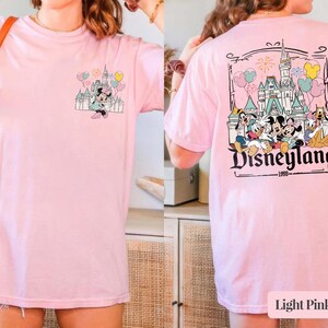 Pode incluir: T-shirt rosa claro com um design da Disneyland na frente e atr&aacute;s. O gr&aacute;fico frontal mostra Minnie Mouse com um castelo e bal&otilde;es. O gr&aacute;fico traseiro mostra personagens da Disneyland e o texto "Disneyland 1955".