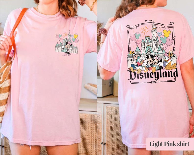 Pode incluir: T-shirt rosa claro com um design da Disneyland na frente e atr&aacute;s. O gr&aacute;fico frontal mostra Minnie Mouse com um castelo e bal&otilde;es. O gr&aacute;fico traseiro mostra personagens da Disneyland e o texto "Disneyland 1955".