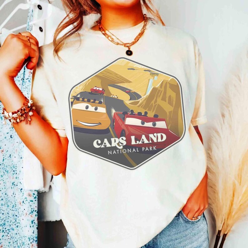 Op de afbeelding: Een cr&egrave;mekleurig t-shirt met een zeshoekige grafische print van een woestijnlandschap met twee antropomorfe auto's. De tekst "CARS LAND NATIONAL PARK" staat in witte letters onderaan de afbeelding, die een weg laat zien.
