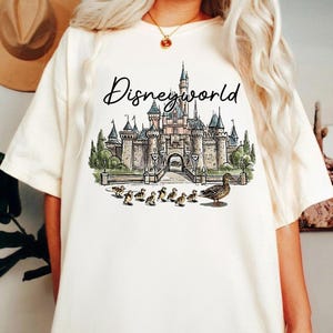 Puede incluir: Camiseta color crema con una ilustraci&oacute;n en acuarela de un castillo y el texto "Disneyworld" encima. La ilustraci&oacute;n incluye una madre pato y patitos. La camiseta es una prenda casual para el d&iacute;a a d&iacute;a.