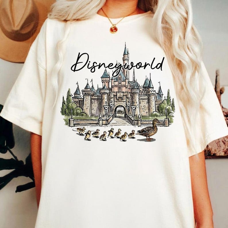 Puede incluir: Camiseta color crema con una ilustraci&oacute;n en acuarela de un castillo y el texto "Disneyworld" encima. La ilustraci&oacute;n incluye una madre pato y patitos. La camiseta es una prenda casual para el d&iacute;a a d&iacute;a.