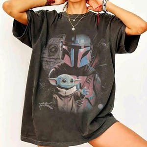 May include: A charcoal gray short-sleeve t-shirt featuring a graphic print of a helmeted character and a small green creature with large ears. The design includes a space station and starships in the background, rendered in a muted color palette.